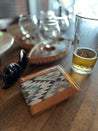 A Drewski Wallet by Zilker Belts, featuring handcrafted leather and Argentine woven fabric, a pair of sunglasses, and a half-full glass of beer sit on a wooden table with empty glassware in the background.