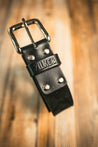 A close-up of the Texas Exes II belt by Zilker Belts, crafted from black Argentine suede leather with a silver buckle and the brand name "ZILKER BELTS" embossed on a loop. The background is a blurred wooden surface.