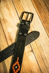 The Texas Exes II by Zilker Belts, a black leather belt with a metallic buckle and an orange and black southwestern pattern, is displayed on a wooden surface, capturing the iconic style favored by Texas Exes.