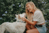 A blonde woman petting a white dog in front of flowers wearing the Zilker Belts Argentina Dog Collar.